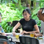 Grupo de adultos reunido em torno de uma mesa, participando de uma discussão em um ambiente arborizado.
