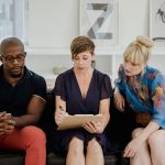 Três adultos conversam seriamente em um ambiente interno, com documentos sobre a mesa.
